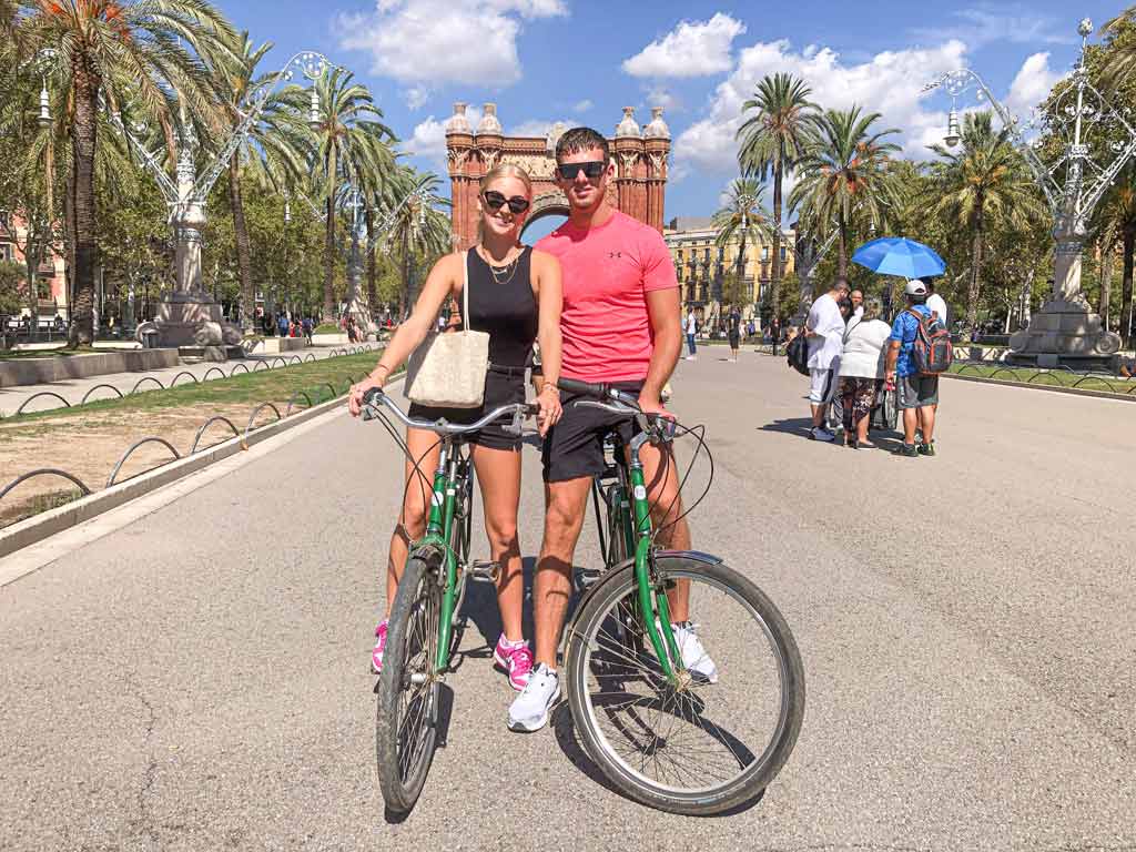 A young couple on a 2-hour private bike tour taking a picture in front of Barcelona's Arc de Triomf, with their bikes parked nearby.