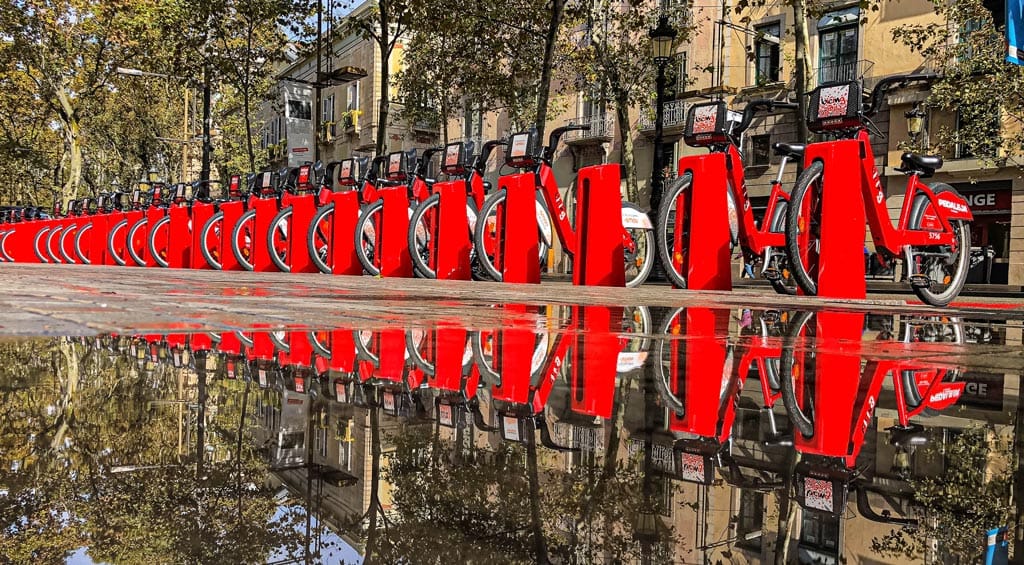Bicing station in Barcelona with multiple red bicycles neatly parked, reflecting in a pool of water on a sunny day.