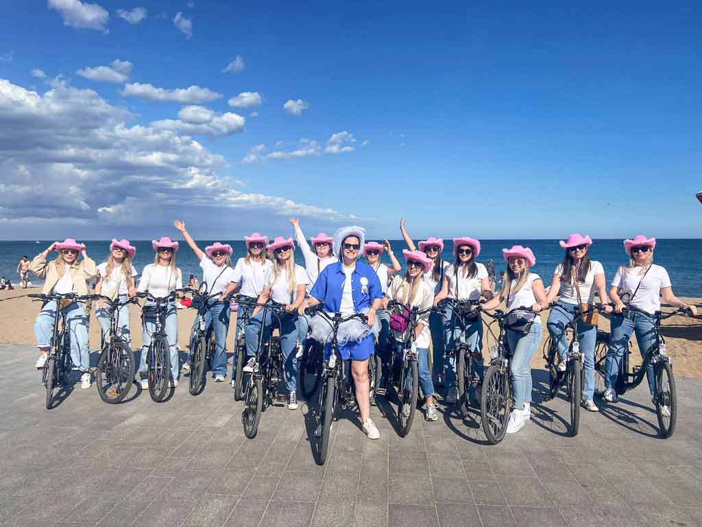 Groep vrouwen op vrijgezellenweekend tijdens een fietstour met Perry Tours, poserend op e-bikes met de zee op de achtergrond