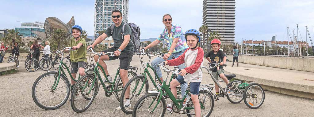 Gezin met drie kinderen poseert bij de haven van Barcelona met fietsen en een aanhangfiets voor het jongste kind.
