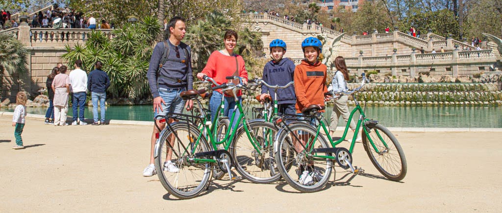 Twee volwassenen en twee kinderen met blauwe fietshelmen poseren met fietsen bij de monumentale fontein in Parc de la Ciutadella.