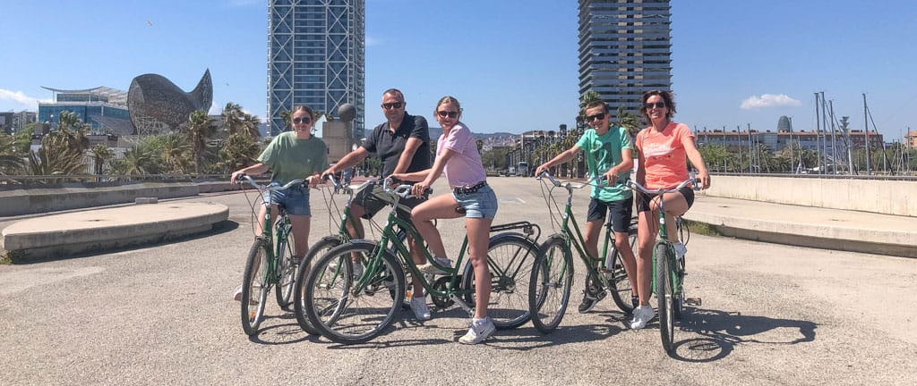 Een gezin van 5 poseren met groene fietsen bij de haven van Barcelona, met de visvormige sculptuur en twee torens op de achtergrond.
