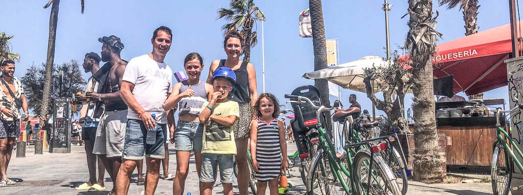 Gezin met drie kinderen neemt een ijsjespauze bij de strandpromenade van Barcelona, naast fietsen met een kinderzitje.