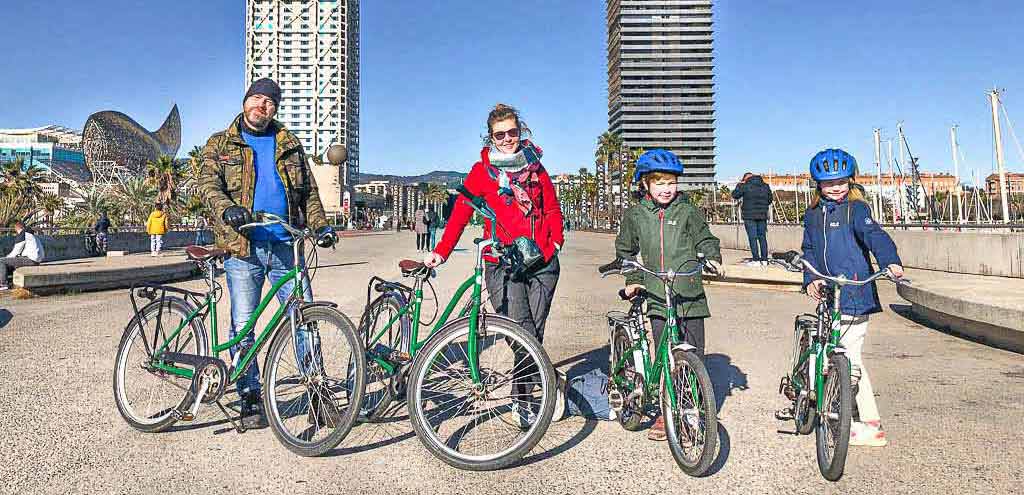 Gezin met twee kinderen met blauwe fietshelmen poseert met fietsen bij de haven van Barcelona.